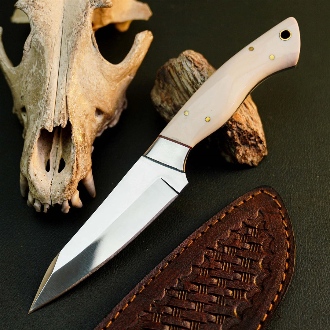 A fixed-blade knife with a polished silver blade and a light beige bone or horn handle, displayed diagonally on a dark textured surface next to a brown leather sheath, a dried animal skull, and a piece of wood.