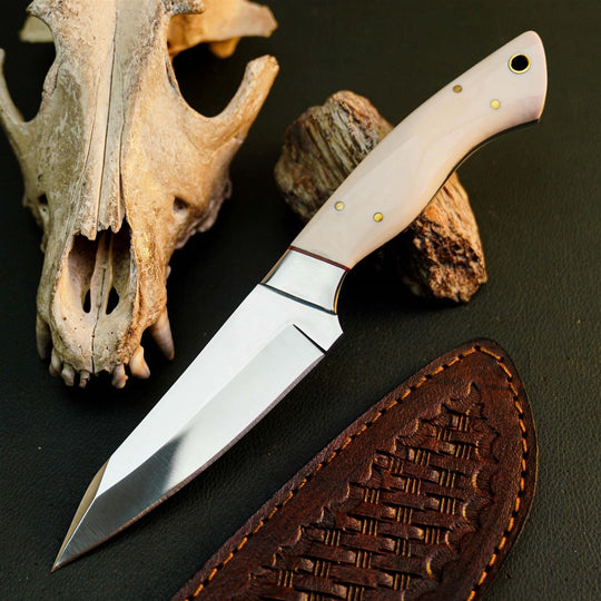 A fixed-blade knife with a polished silver blade and a light beige bone or horn handle, displayed diagonally on a dark textured surface next to a brown leather sheath, a dried animal skull, and a piece of wood.
