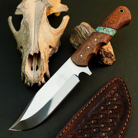A fixed-blade knife with a polished silver blade, a brown wood handle featuring a turquoise inlay, and a brown woven leather sheath, arranged on a dark textured surface with a bone skull and wood props.