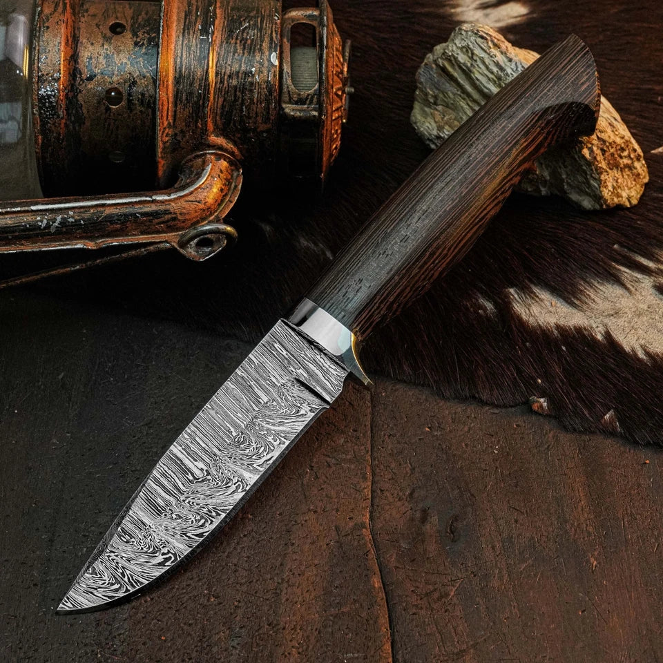 A fixed-blade knife with a patterned Damascus steel blade and a dark wood handle, displayed diagonally on a rustic dark wooden surface with a cowhide rug, a rock, and a vintage lantern in the background.