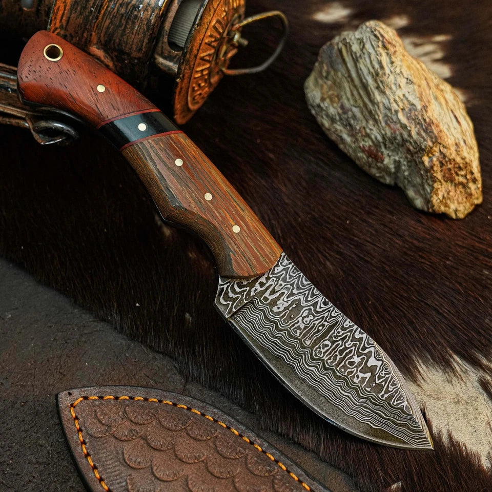 A fixed-blade knife with a patterned Damascus steel blade and a reddish-brown wooden handle, displayed diagonally on dark brown fur with a leather sheath and rustic stone props.