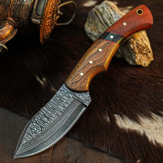 A fixed-blade knife with a patterned Damascus steel blade and a reddish-brown wooden handle, displayed diagonally on a dark brown and white cowhide rug with rustic stones and a metal container in the background.