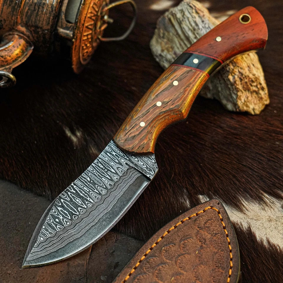A fixed-blade knife with a patterned Damascus-style steel blade and a contoured wooden handle, displayed diagonally on a dark fur surface with a leather sheath and rustic props in the background.