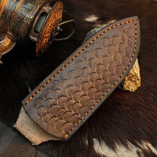 A close-up of a dark brown leather knife sheath with an embossed scallop pattern and contrast stitching, resting on a cowhide surface next to a rustic wooden object.