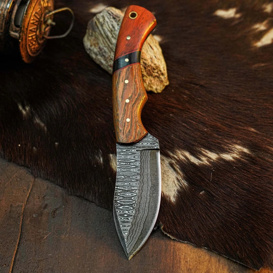 A fixed-blade knife with a patterned Damascus steel blade and a reddish-brown wood handle, displayed on a dark brown and white cowhide rug with rustic props.