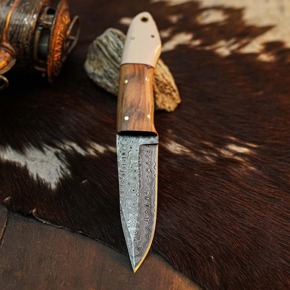 A custom handmade knife with a patterned Damascus steel blade, a wooden handle, and a white end cap, displayed on a brown and white cowhide rug.