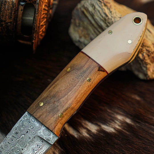 A close-up of a handcrafted knife with a Damascus steel blade and a wooden handle featuring a light-colored inlay, resting on a dark textured surface with rustic wood props.