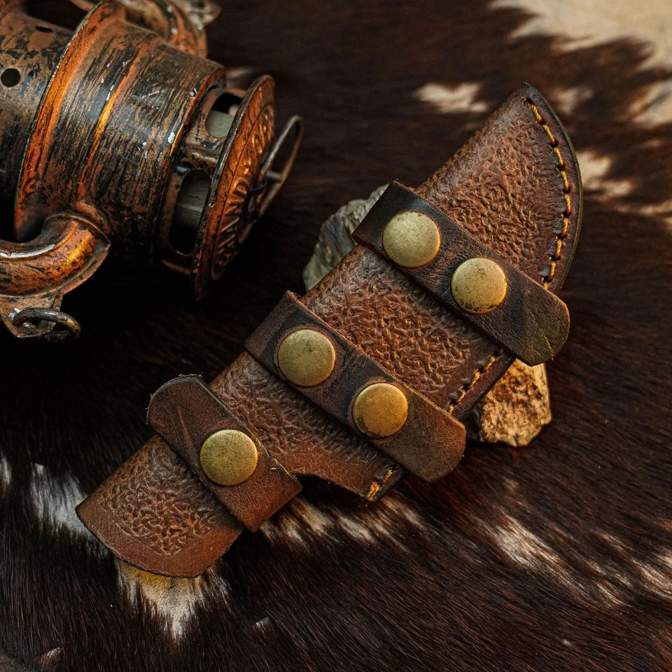 A close-up of a brown leather knife sheath with brass-tone rivets and strap details, placed on a cowhide surface next to a rustic metal object.