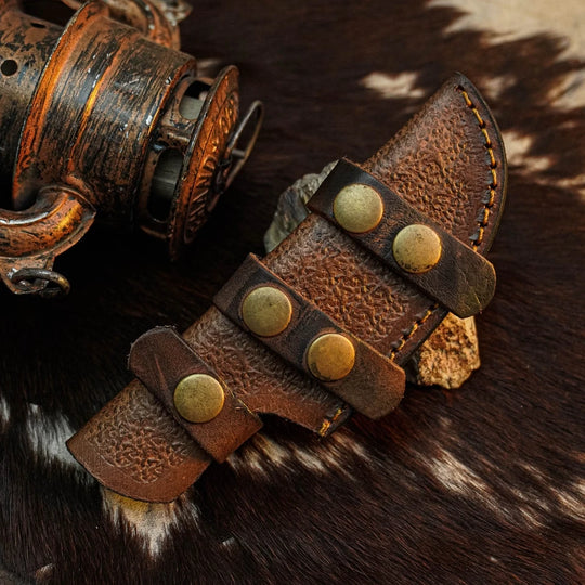 A close-up of a brown leather knife sheath with brass-tone rivets and strap details, placed on a cowhide surface next to a rustic metal object.