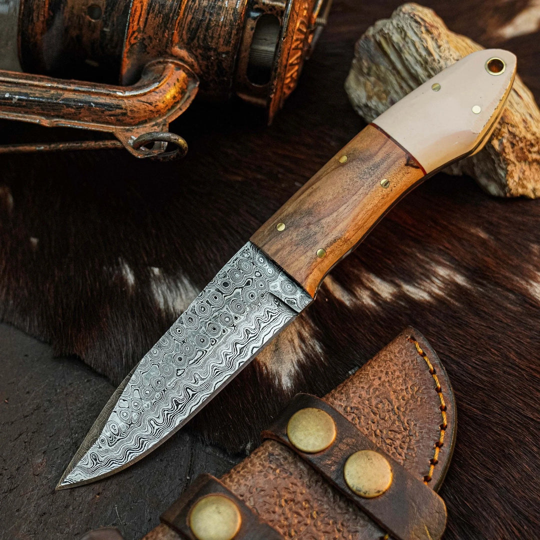 A fixed-blade knife with a patterned Damascus steel blade, a brown wood handle, and a cream-colored end piece, displayed diagonally on a dark textured surface with a leather sheath and rustic props.