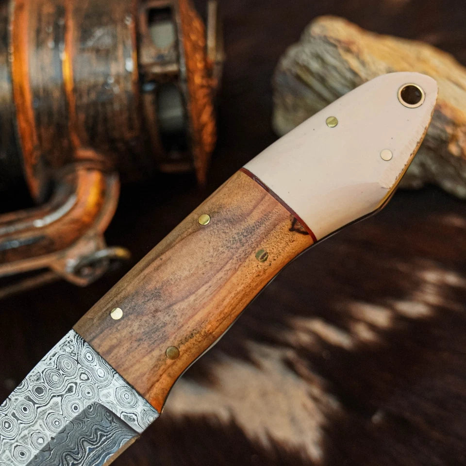A close-up of a handcrafted knife with a Damascus steel blade, a brown wood handle, and brass pins, resting on a dark textured surface with rustic props in the background.