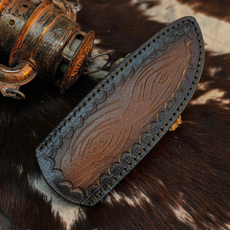 A brown leather knife sheath with embossed floral and scroll patterns, featuring decorative stitching along the curved edge, displayed on a cowhide surface next to a rustic metal object.