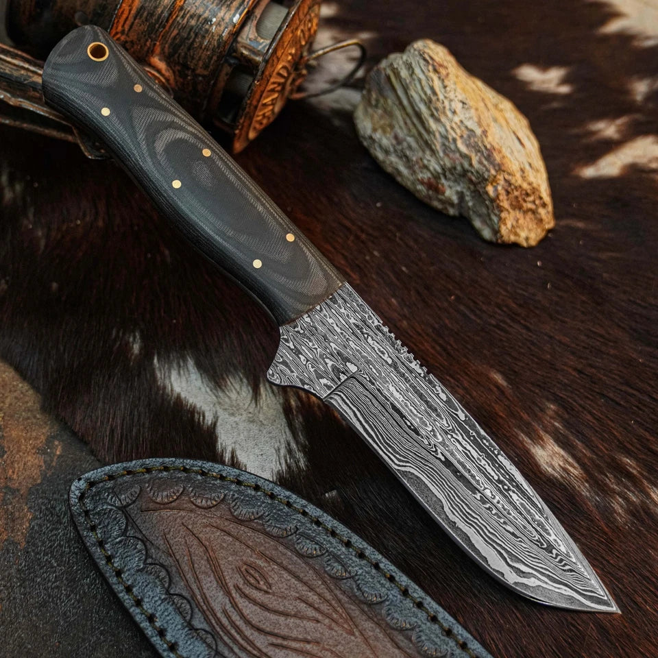 A fixed-blade knife with a patterned Damascus-style blade and dark handle, styled on a brown and white cowhide surface with a leather sheath, wooden object, and rock nearby.