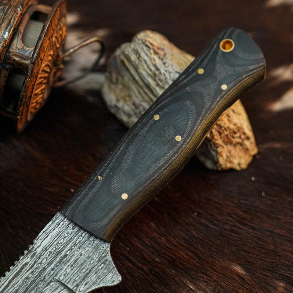A close-up of a fixed-blade knife with a patterned steel blade and a dark textured handle, resting on rocks over a dark wooden surface.
