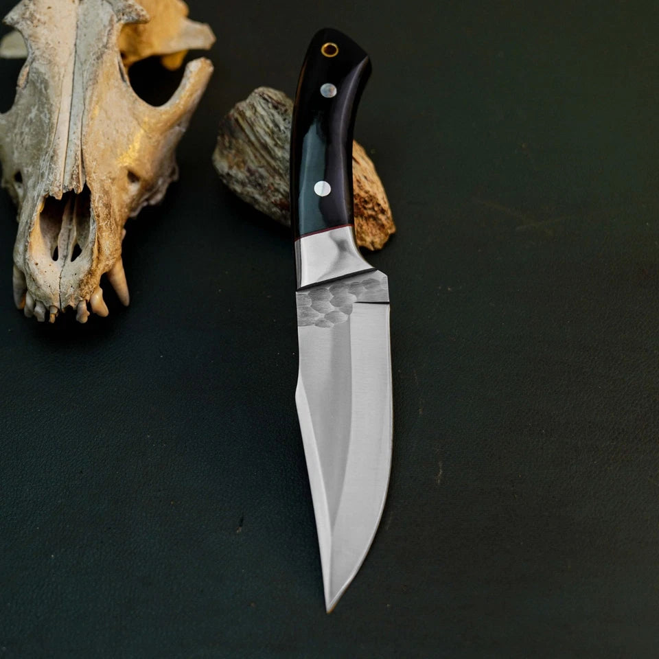 A fixed-blade knife with a hammered silver blade and a black handle with silver rivets, displayed on a dark textured surface next to animal skull and bone props.