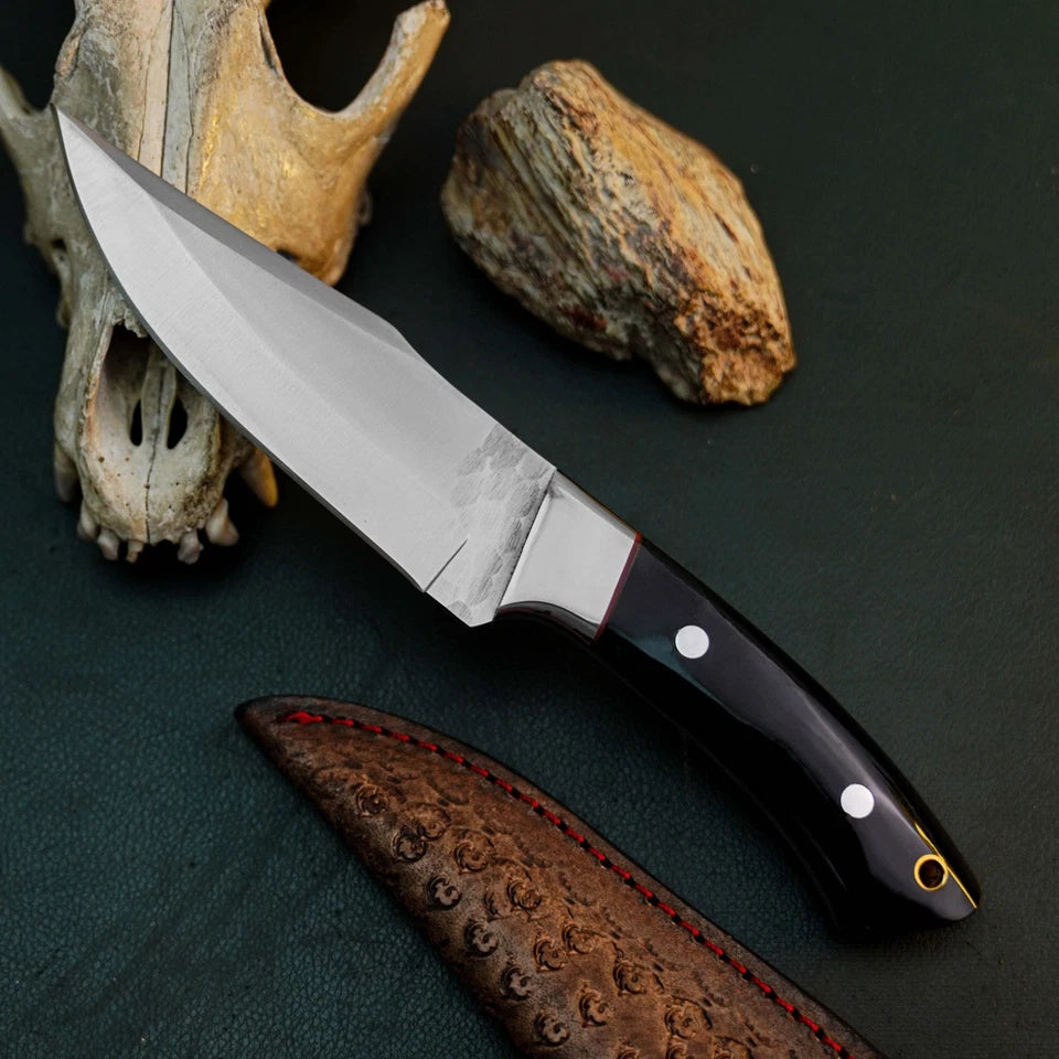 A fixed-blade knife with a satin-finished silver blade and a black handle with silver rivets, displayed diagonally on a dark textured surface next to its brown textured sheath. The background is styled with animal bones and a rock.