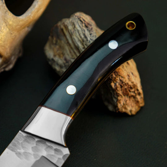 A close-up of a knife with a dark glossy handle and hammered metal blade, resting on rocks against a dark background.