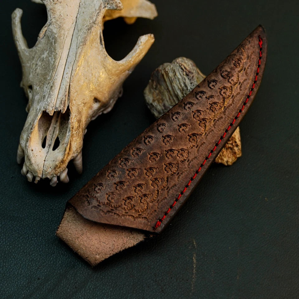 A dark brown leather knife sheath with a textured, embossed pattern and red stitching, displayed next to a carved wooden block and an animal skull on a dark background.