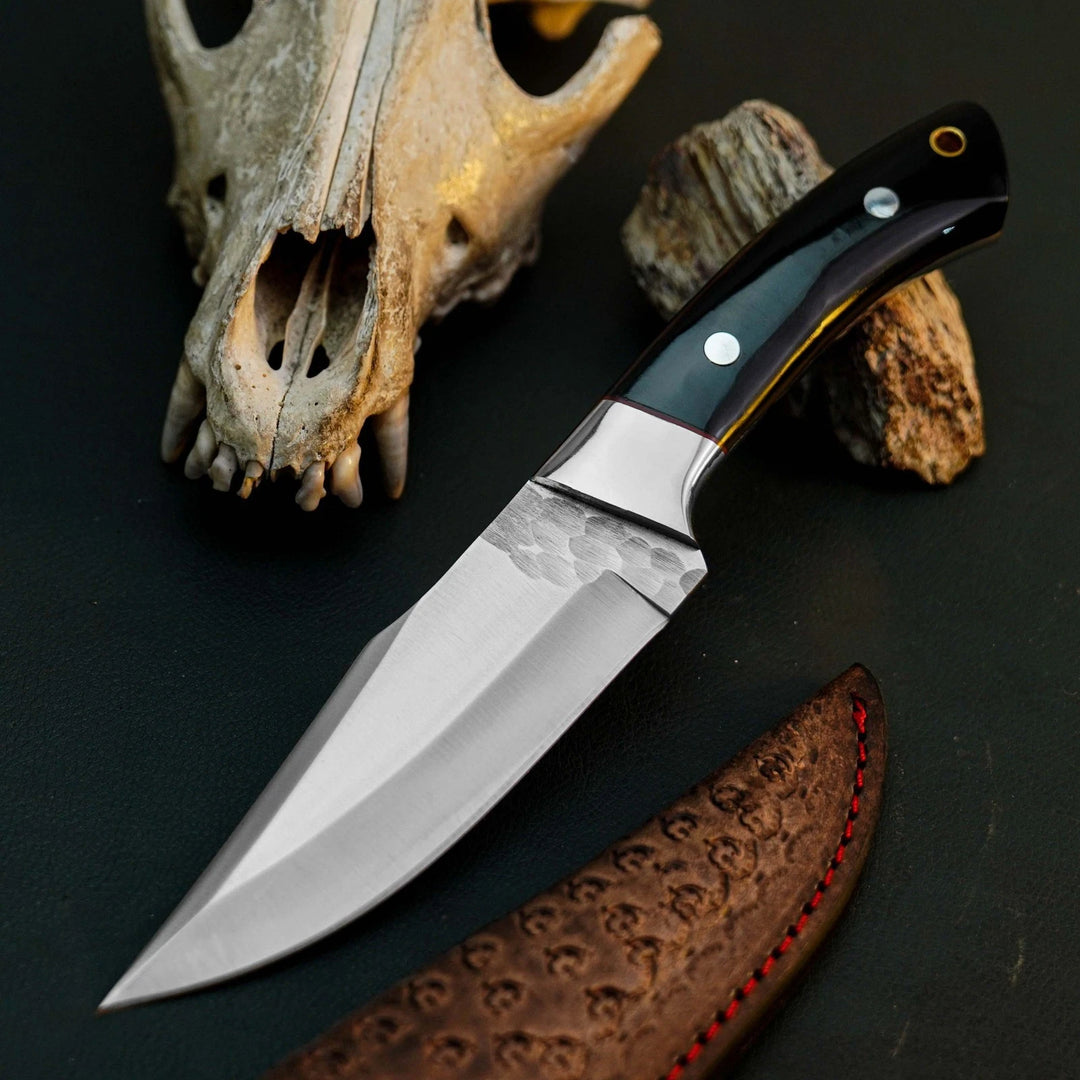 A fixed-blade knife with a hammered steel blade and a dark handle lies on a dark surface, styled with a bone skull, wood pieces, and part of a brown leather sheath with red stitching.