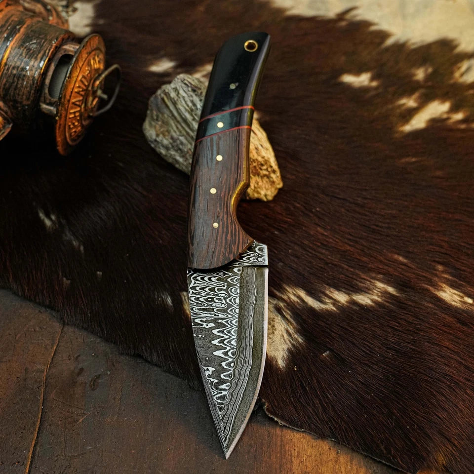 A fixed-blade knife with a Damascus steel pattern blade and a dark wood handle, displayed on a brown and white cowhide rug with rustic props.
