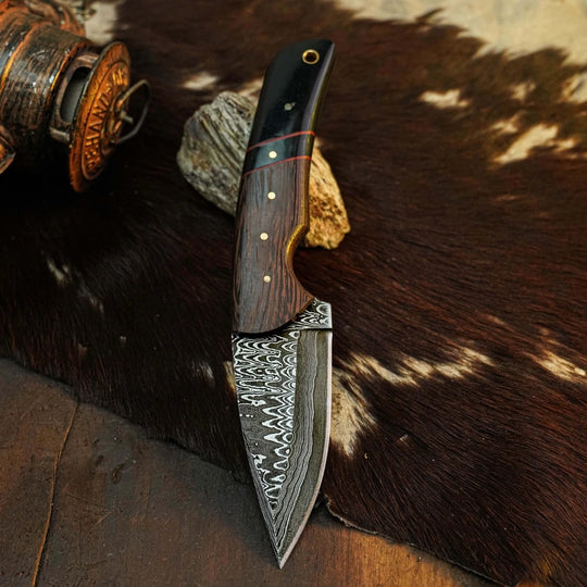 A fixed-blade knife with a Damascus steel pattern blade and a dark wood handle, displayed on a brown and white cowhide rug with rustic props.