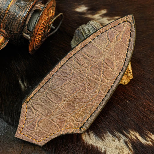A close-up of a brown leather knife sheath with visible stitching, placed on a dark brown and white cowhide surface with rustic metal objects in the background.