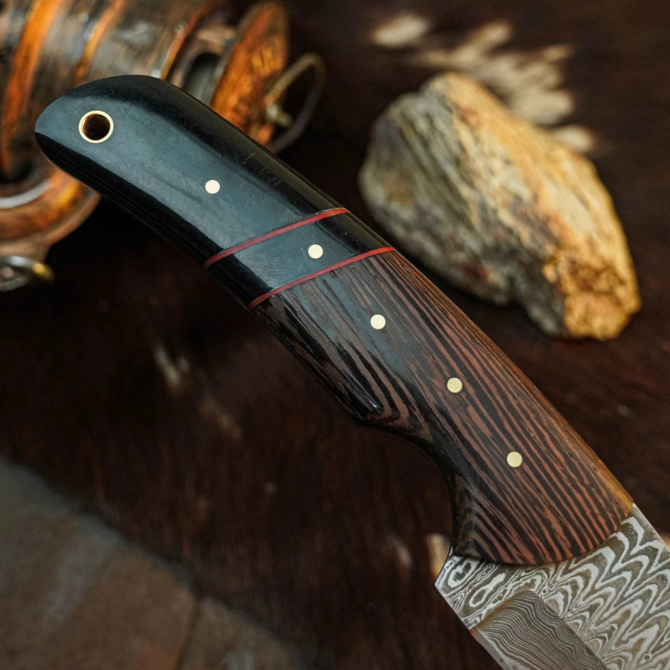 A close-up of a Damascus steel knife with a dark wood handle featuring brass pins and red inlay lines, resting on a rustic wooden surface with a stone and copper-toned object in the background.