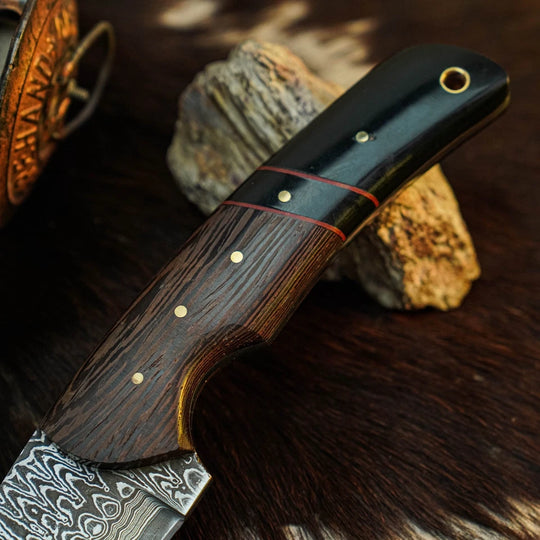 A close-up of a handcrafted knife with a Damascus steel blade, a dark wood handle, and a black end section, resting on a textured brown fur surface with rocks in the background.