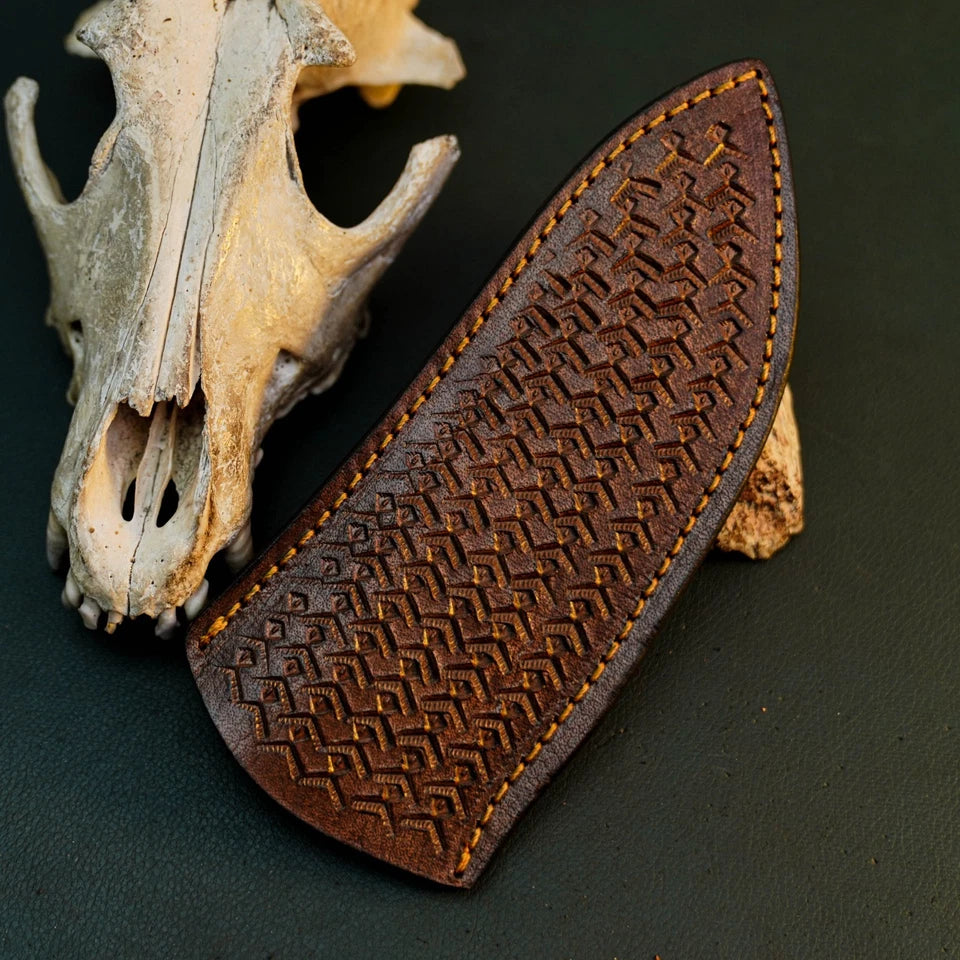 A dark brown leather knife sheath with an embossed geometric pattern and yellow stitching, displayed next to a weathered animal skull on a dark background.