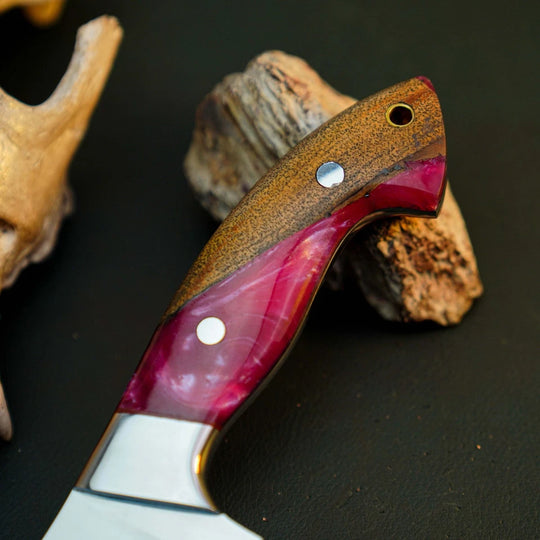 A close-up of a knife handle with a brown wood top section and a magenta-pink resin inlay section, secured with silver pins, resting on driftwood against a dark background.