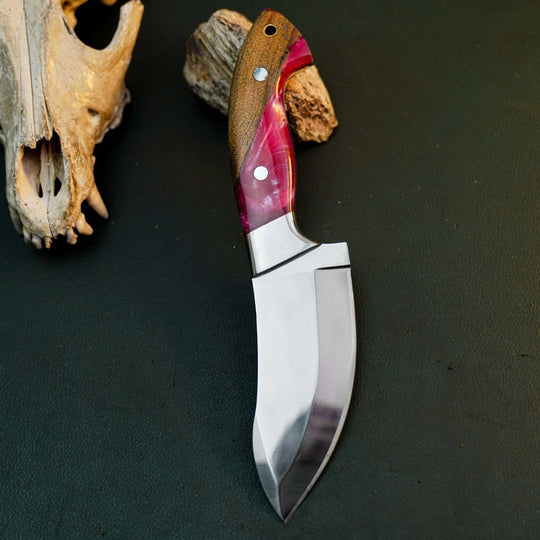 A fixed-blade knife with a polished silver blade and a brown wood handle featuring pink and purple inlay, displayed on a dark textured surface next to a bone skull prop.