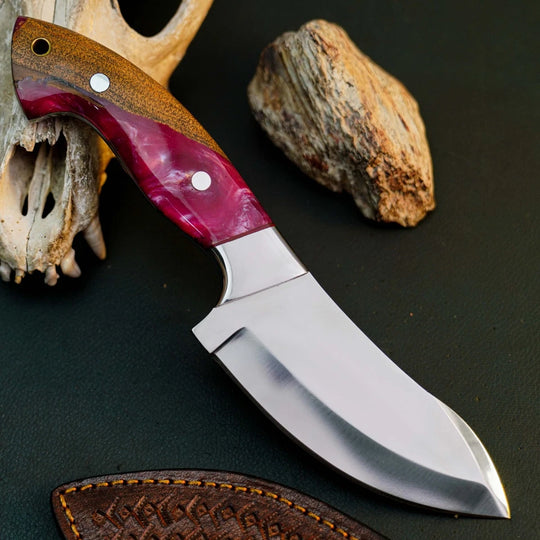 A fixed-blade knife with a polished silver steel blade and a marbled wood handle in brown and deep magenta, displayed diagonally on a dark textured surface with a leather sheath, a bone fragment, and a rock nearby.