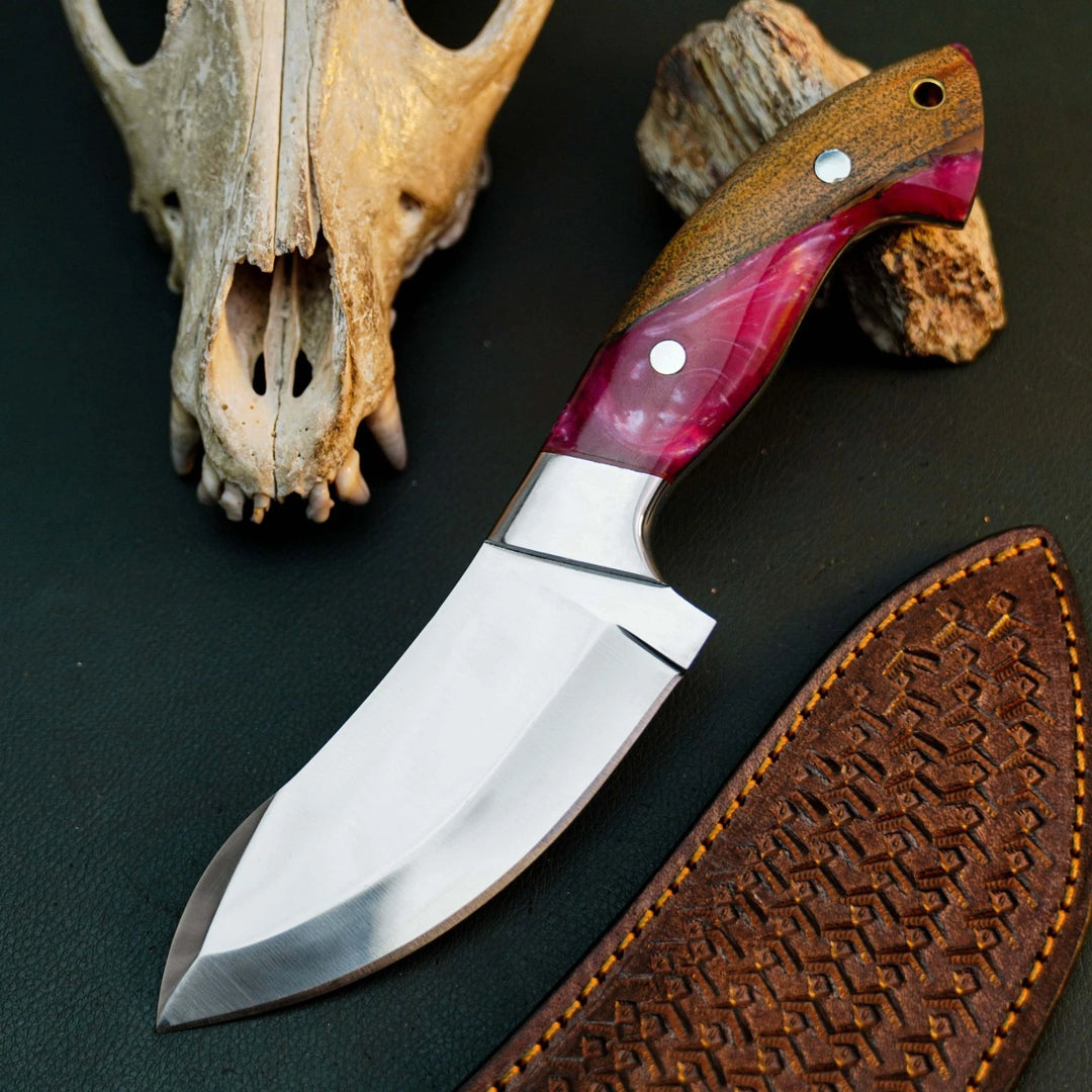 A fixed-blade knife with a polished silver blade, a brown wood handle with a pink inlay, and a brown leather sheath, arranged on a dark surface with a bone skull and wood prop in the background.